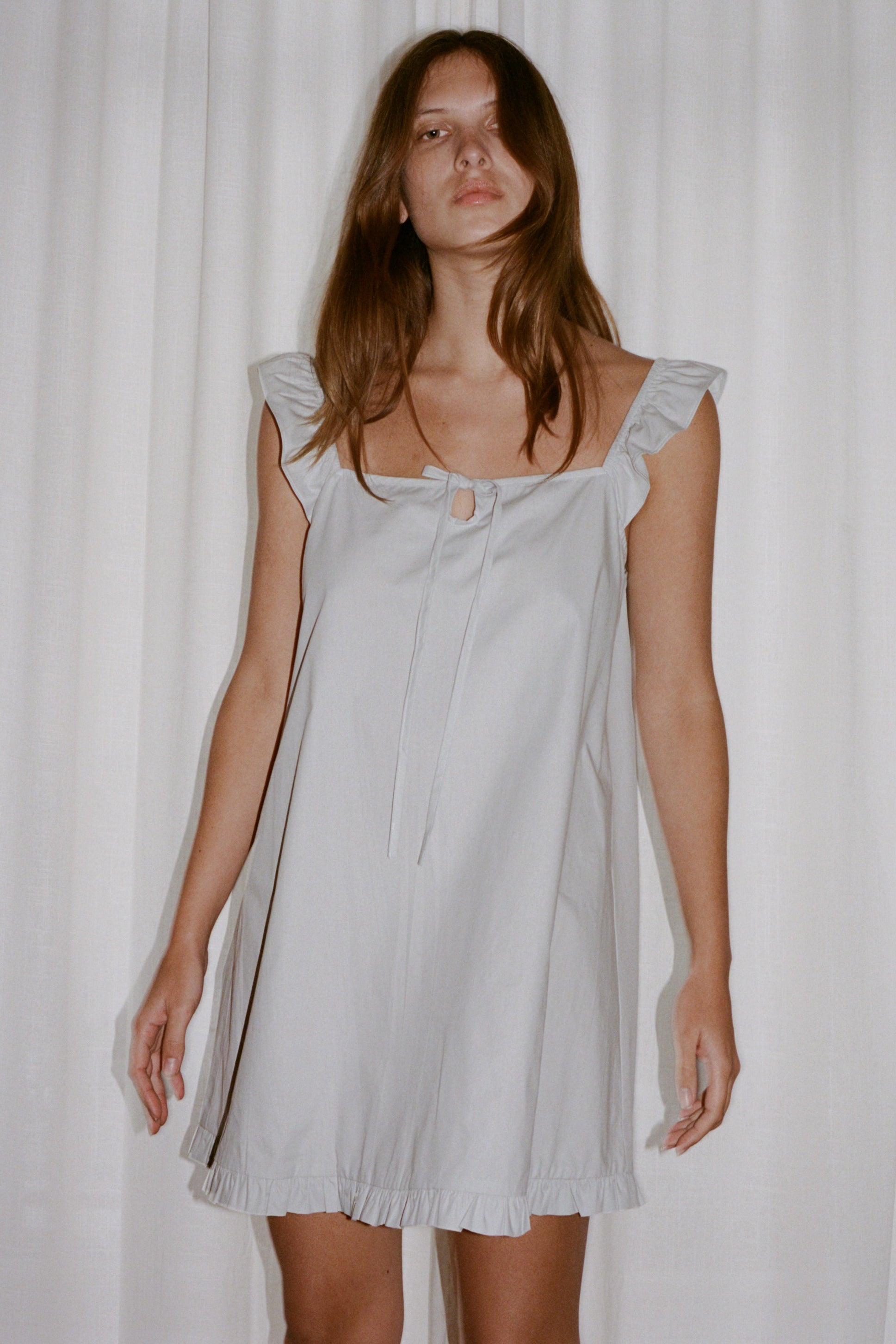 A woman with long brown hair stands by white curtains, wearing the Jonni Dress - Powder Blue by 0. This organic cotton dress has ruffled sleeves and hem, plus a keyhole opening with a tie at the neckline.