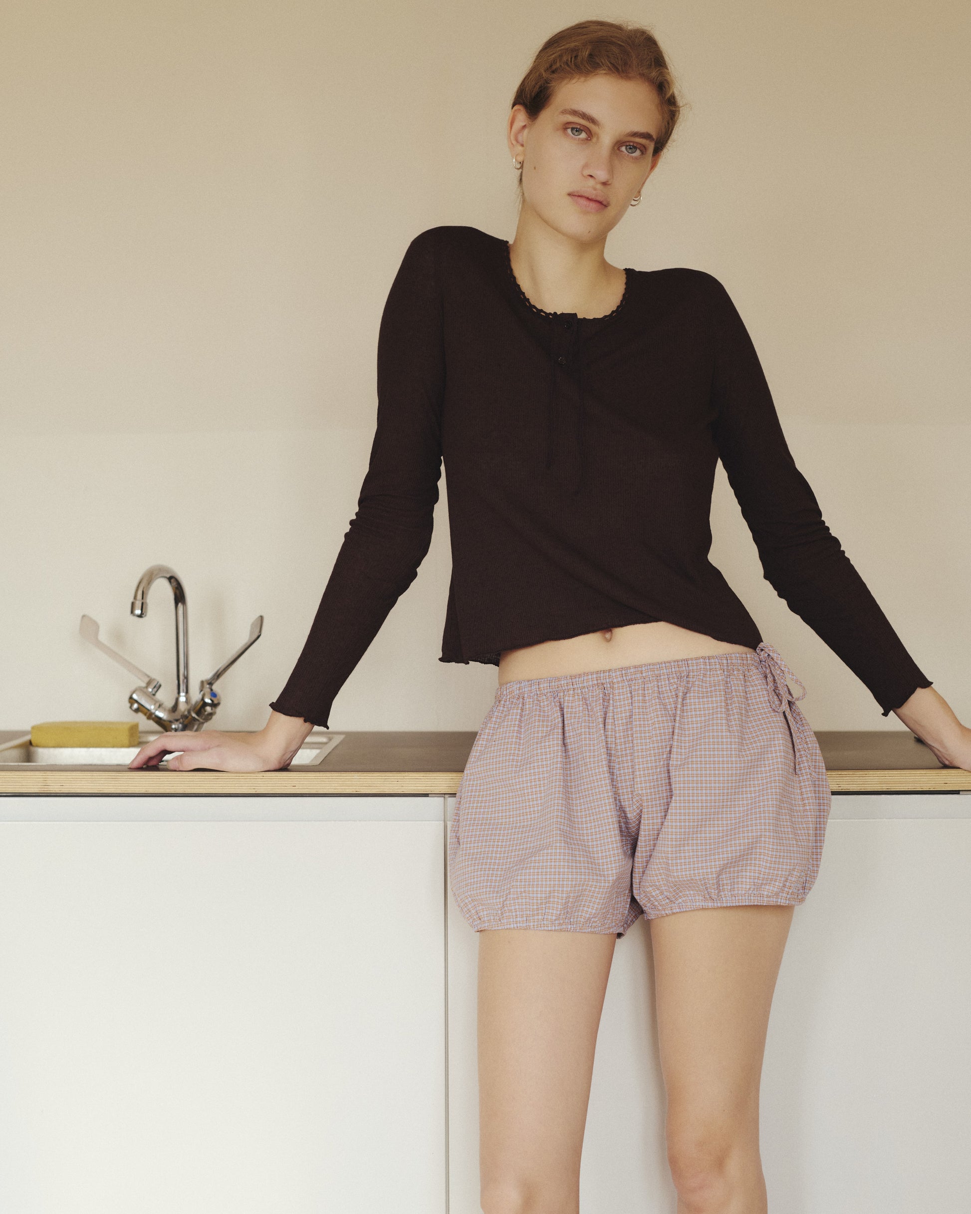 Wearing the Rosa Long Sleeve in Chocolate by 0—a soft modal-linen blend top with organic cotton trim—and checked shorts, a person with light hair leans against a kitchen counter beside a sink, gazing at the camera with a neutral expression.