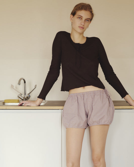 Wearing the Rosa Long Sleeve in Chocolate by 0—a soft modal-linen blend top with organic cotton trim—and checked shorts, a person with light hair leans against a kitchen counter beside a sink, gazing at the camera with a neutral expression.