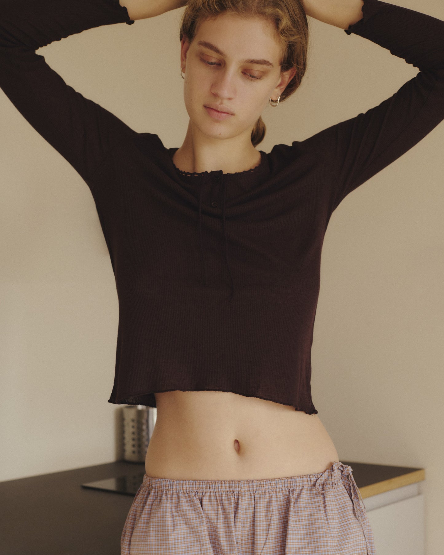 A person with short hair stands indoors, wearing the 0 Rosa Long Sleeve in Chocolate, which features an organic cotton trim, paired with plaid pajama pants. Their arms are raised behind their head as they look down with a calm expression.