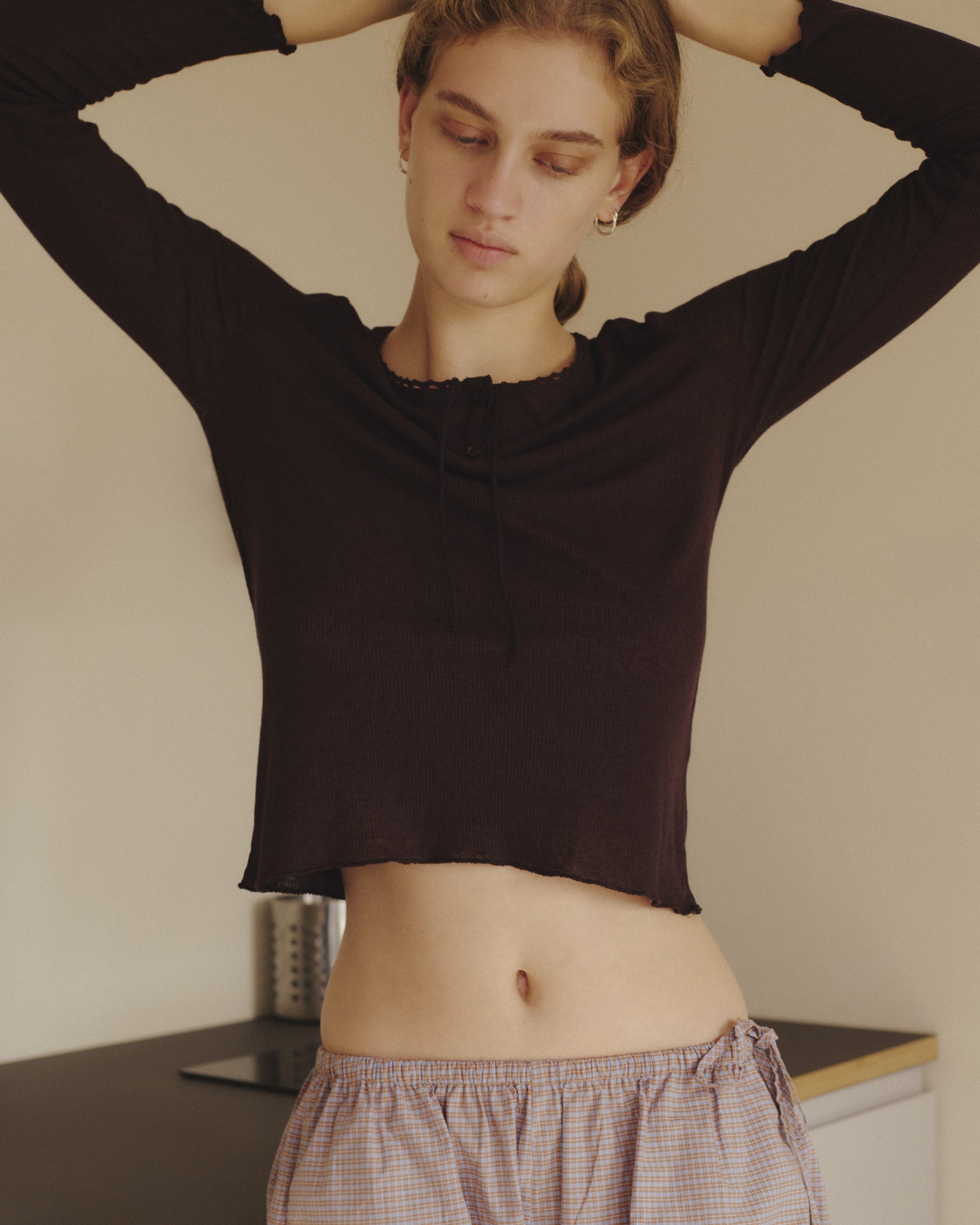 A person with short hair stands indoors, wearing the 0 Rosa Long Sleeve in Chocolate, which features an organic cotton trim, paired with plaid pajama pants. Their arms are raised behind their head as they look down with a calm expression.