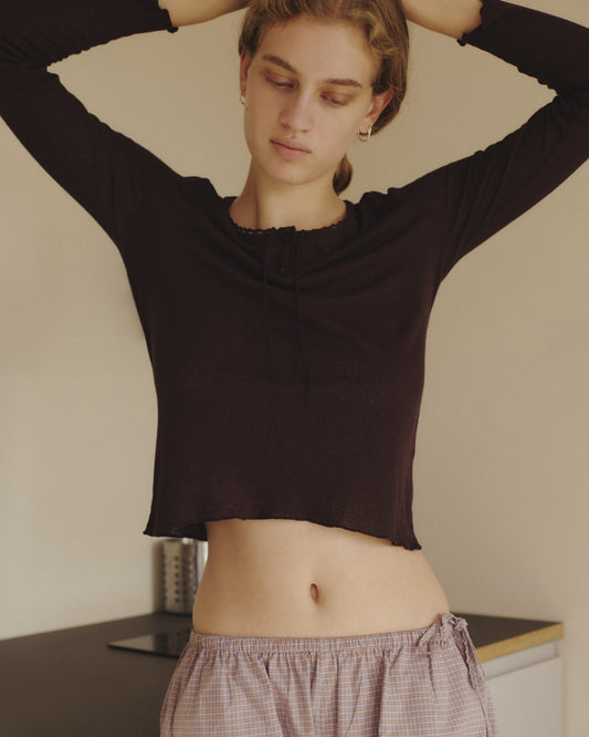 A person with short hair stands indoors, wearing the 0 Rosa Long Sleeve in Chocolate, which features an organic cotton trim, paired with plaid pajama pants. Their arms are raised behind their head as they look down with a calm expression.