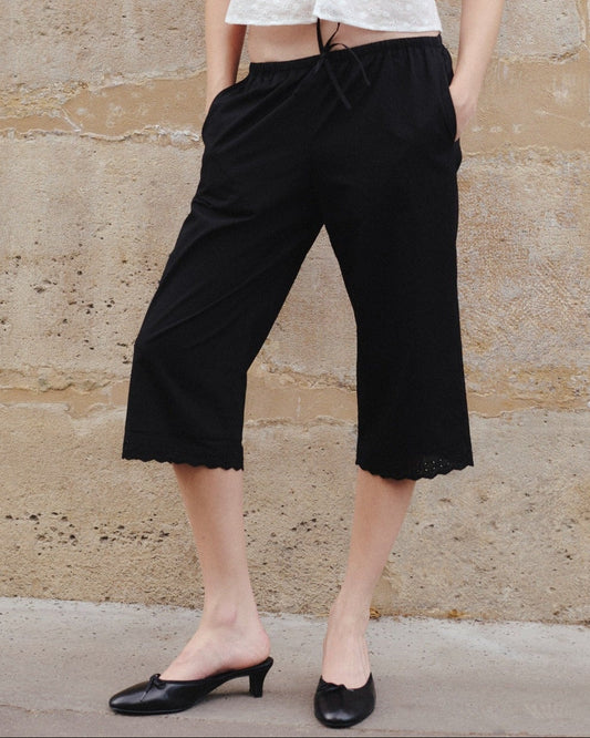 A person wearing the 0 Ellery Capri in black, styled with a white broderie anglaise top and black low-heeled shoes, stands against a textured stone wall with hands in pockets.
