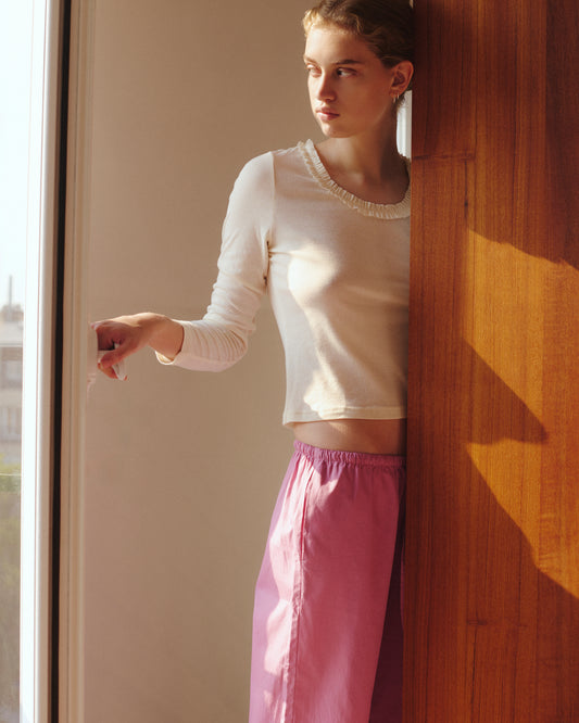 Wearing the Ellery Capri in Mauve by 0 and a white long-sleeve top, a person stands by a wooden wall, gazing out of a window as soft sunlight fills the scene.