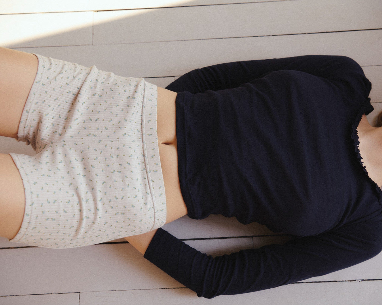 A person is lying on a wooden floor, wearing a dark long-sleeve shirt and the Maggie Short - Blue Floral by Juem, made from light-colored organic cotton with a small pattern. Their face is not visible in the image.
