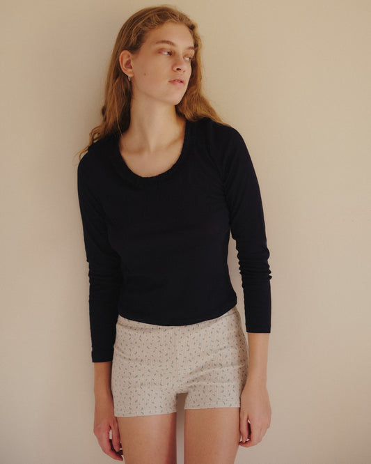 A person with long, wavy hair wears a black long-sleeve top and the Maggie Short - Blue Floral by Juem, standing against a plain beige wall and looking to the side.