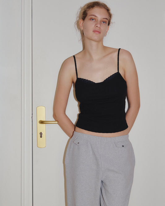 A person stands in front of a white door, wearing the 0 Joan Tank in black with adjustable straps and gray sweatpants, hands behind their back and a neutral expression.