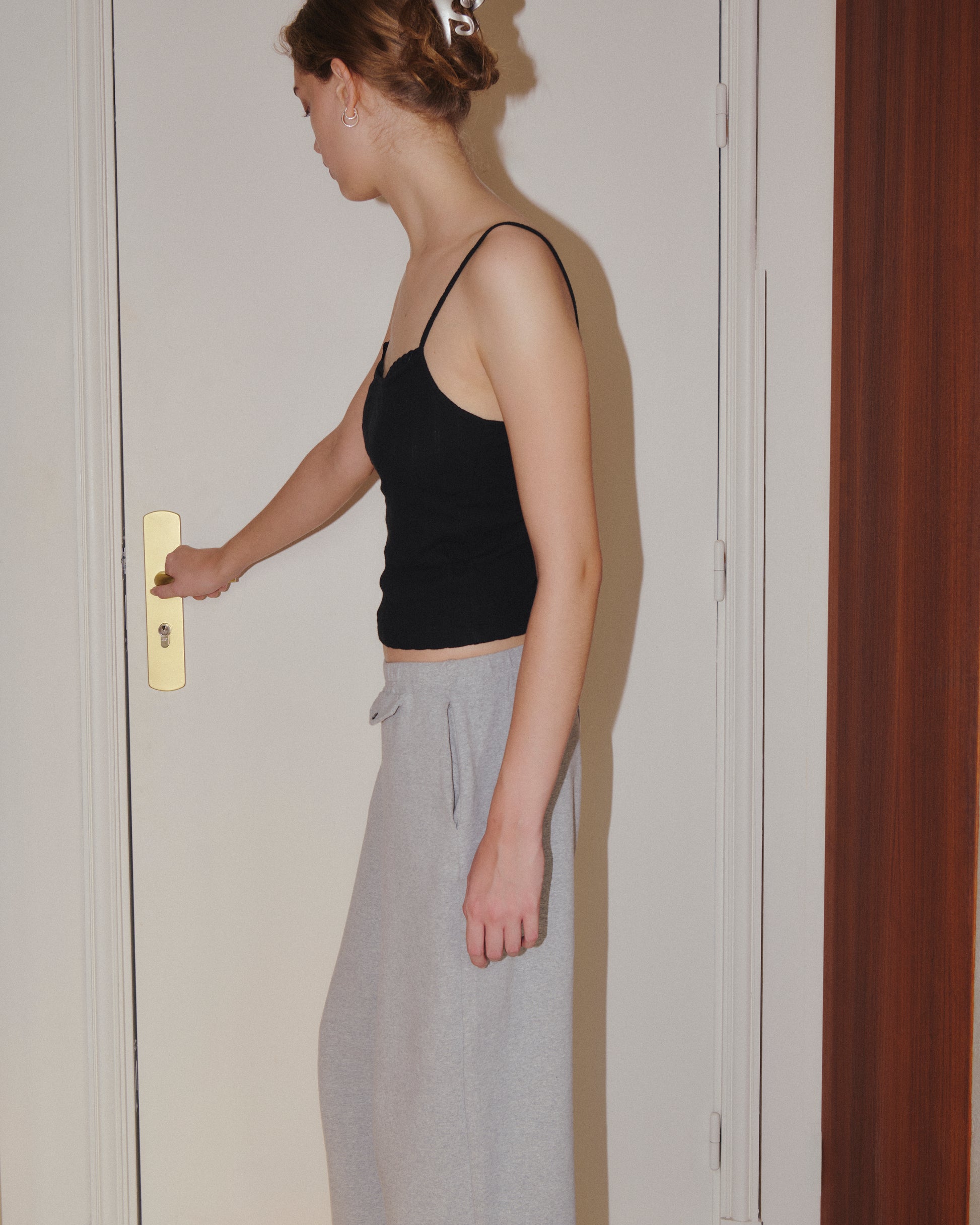 Wearing the Joan Tank - Black by 0, a woman in gray sweatpants reaches for a door handle, standing next to a white door with a gold-colored lock.