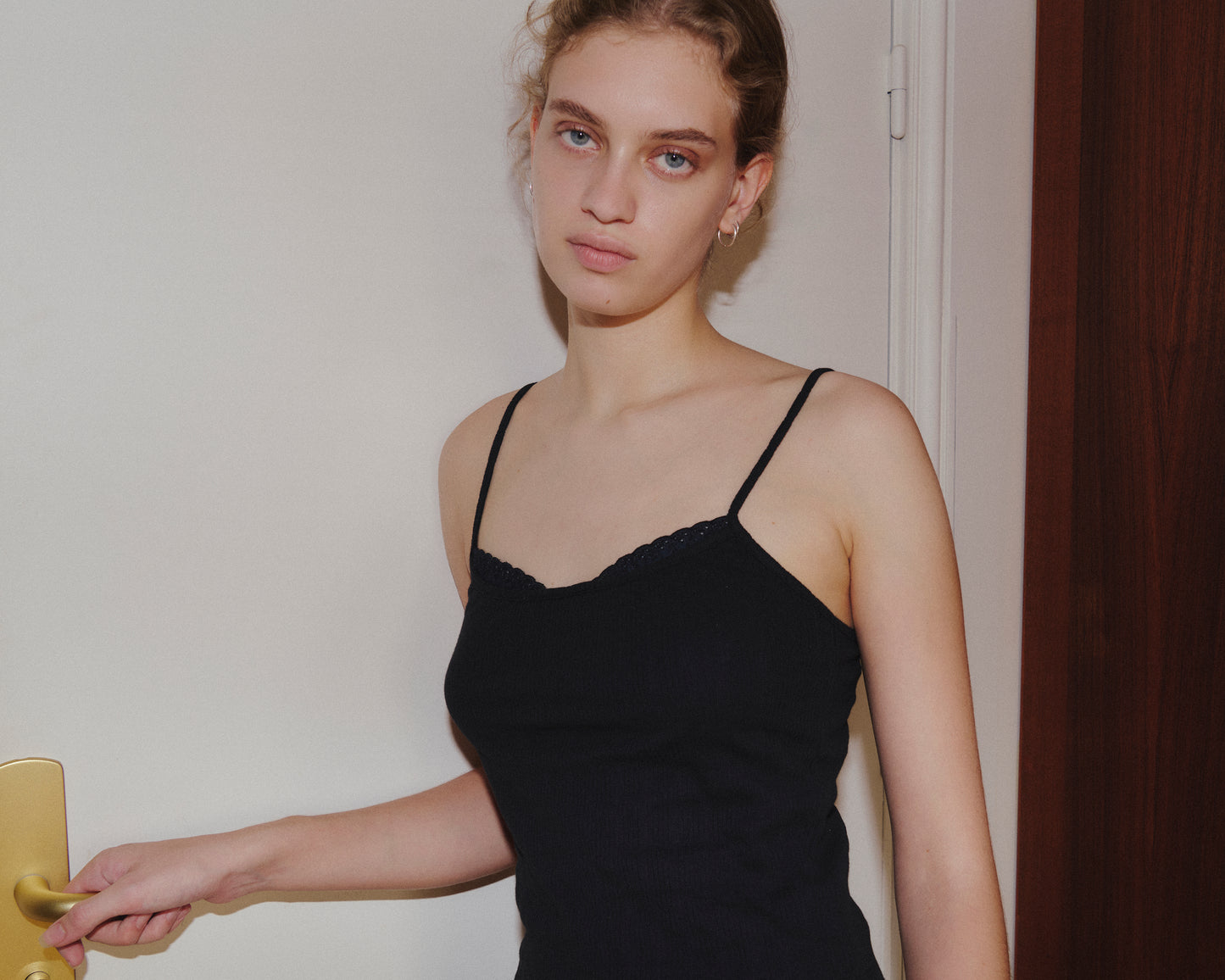 Wearing the 0 Joan Tank in Black, a young woman with curly hair stands indoors, calmly looking at the camera while holding a door handle.