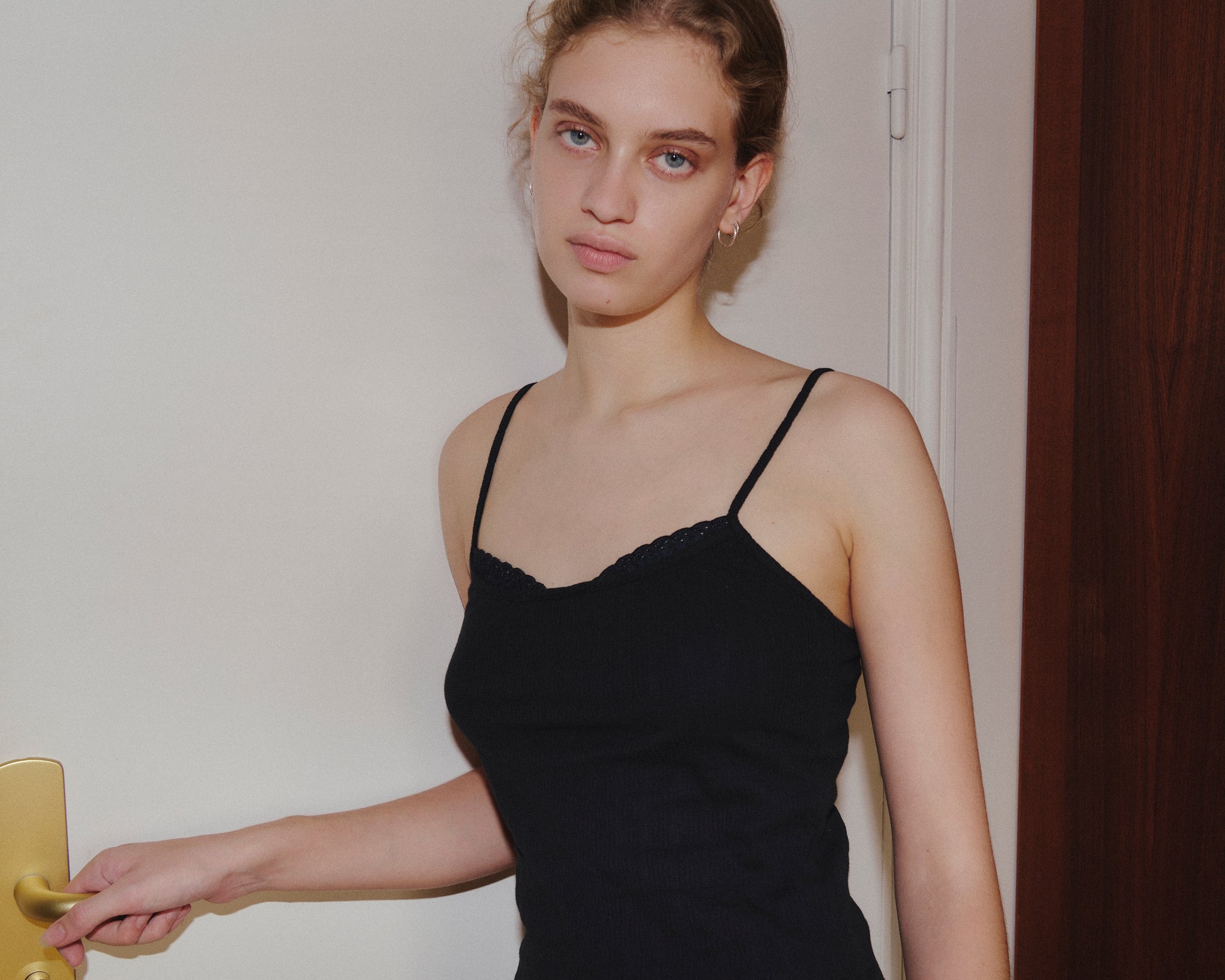 Wearing the 0 Joan Tank in Black, a young woman with curly hair stands indoors, calmly looking at the camera while holding a door handle.
