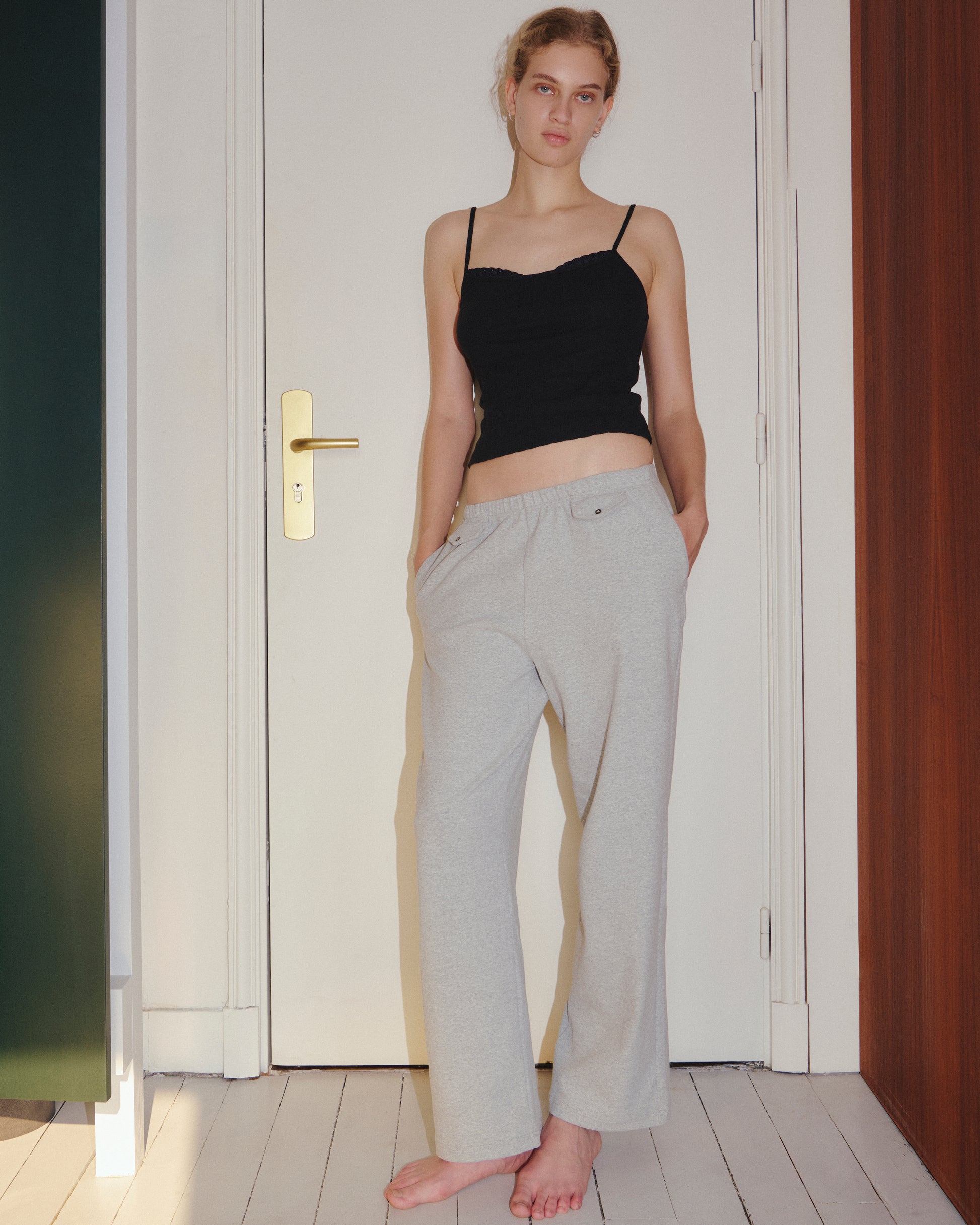 A woman stands barefoot indoors, wearing the 0 Joan Tank - Black with adjustable spaghetti straps and light gray pants, her hands in her pockets, in front of a closed white door.