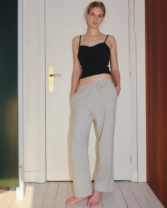 A woman stands barefoot indoors, wearing the 0 Joan Tank - Black with adjustable spaghetti straps and light gray pants, her hands in her pockets, in front of a closed white door.