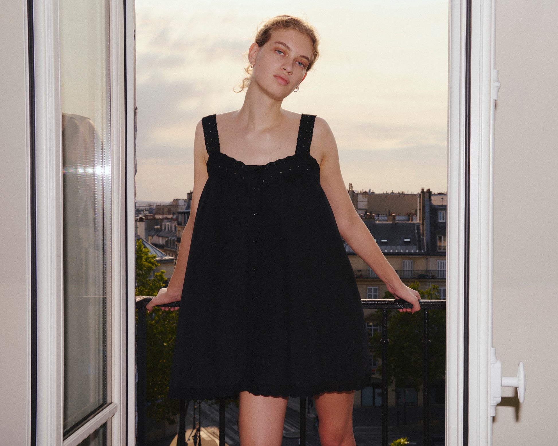 Wearing the Ottie Dress - Black by 0, a woman stands in a doorway at sunset, leaning on a balcony railing with a cityscape in the background.