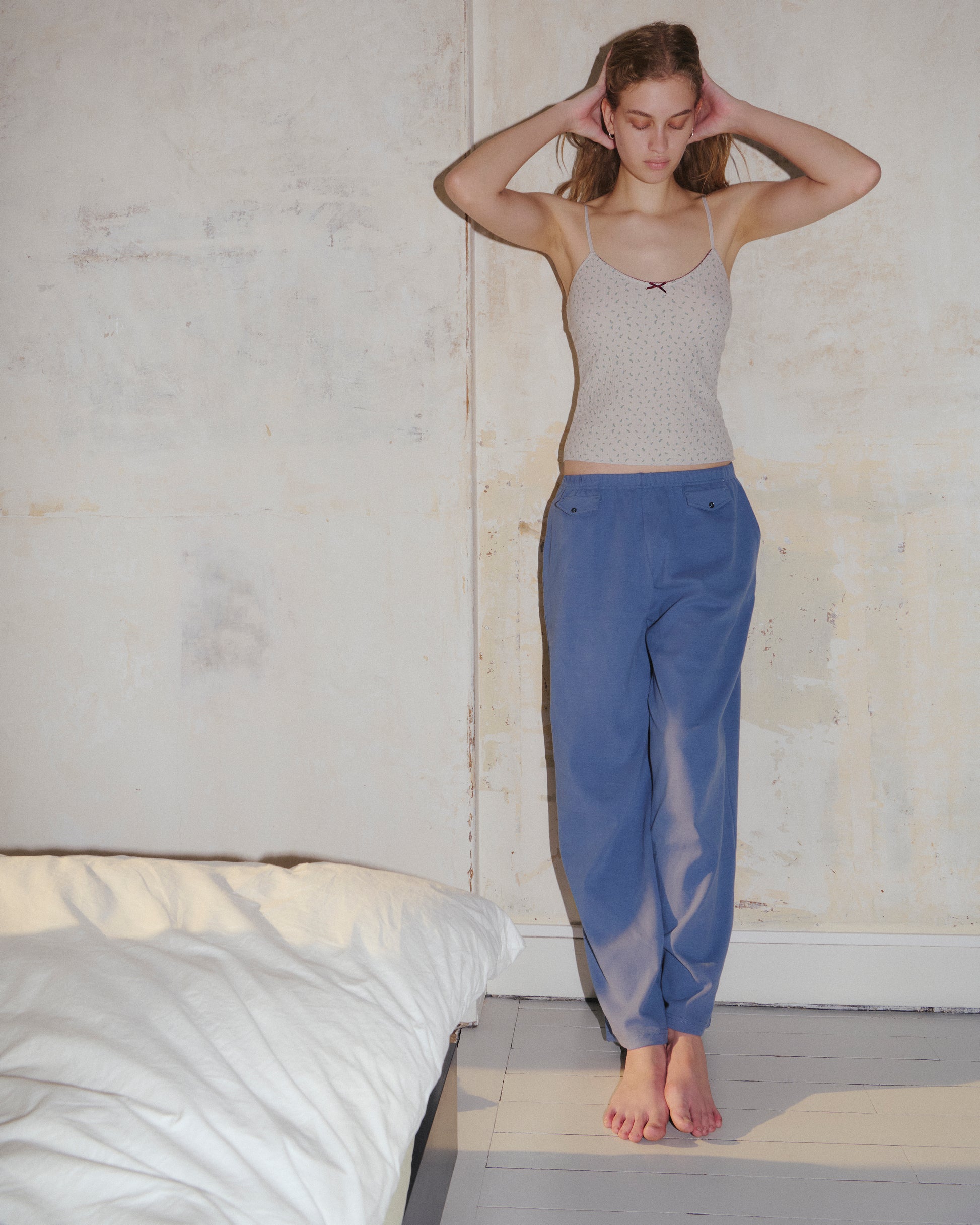 A person stands barefoot on a wooden floor beside a bed, wearing a beige tank top and the Vera Track Pant in Sea Blue by 0, featuring a relaxed fit and elasticated waistband, hands resting behind their head against a light, textured wall.