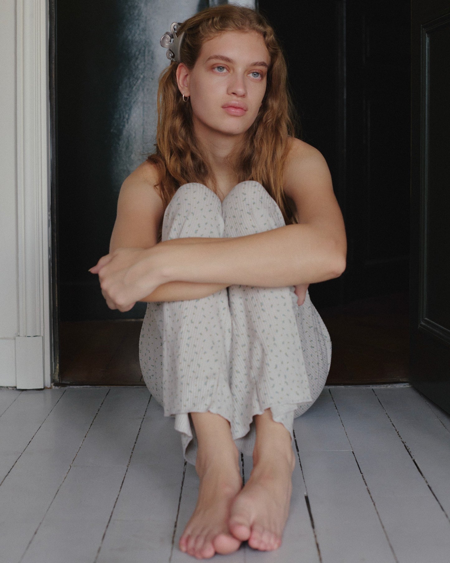A young woman with light brown hair sits on a white wooden floor, hugging her knees to her chest. She wears the Juem Maya Pant in Blue Floral—light-colored pajamas made from comfortable, organic cotton—and looks thoughtfully into the distance.