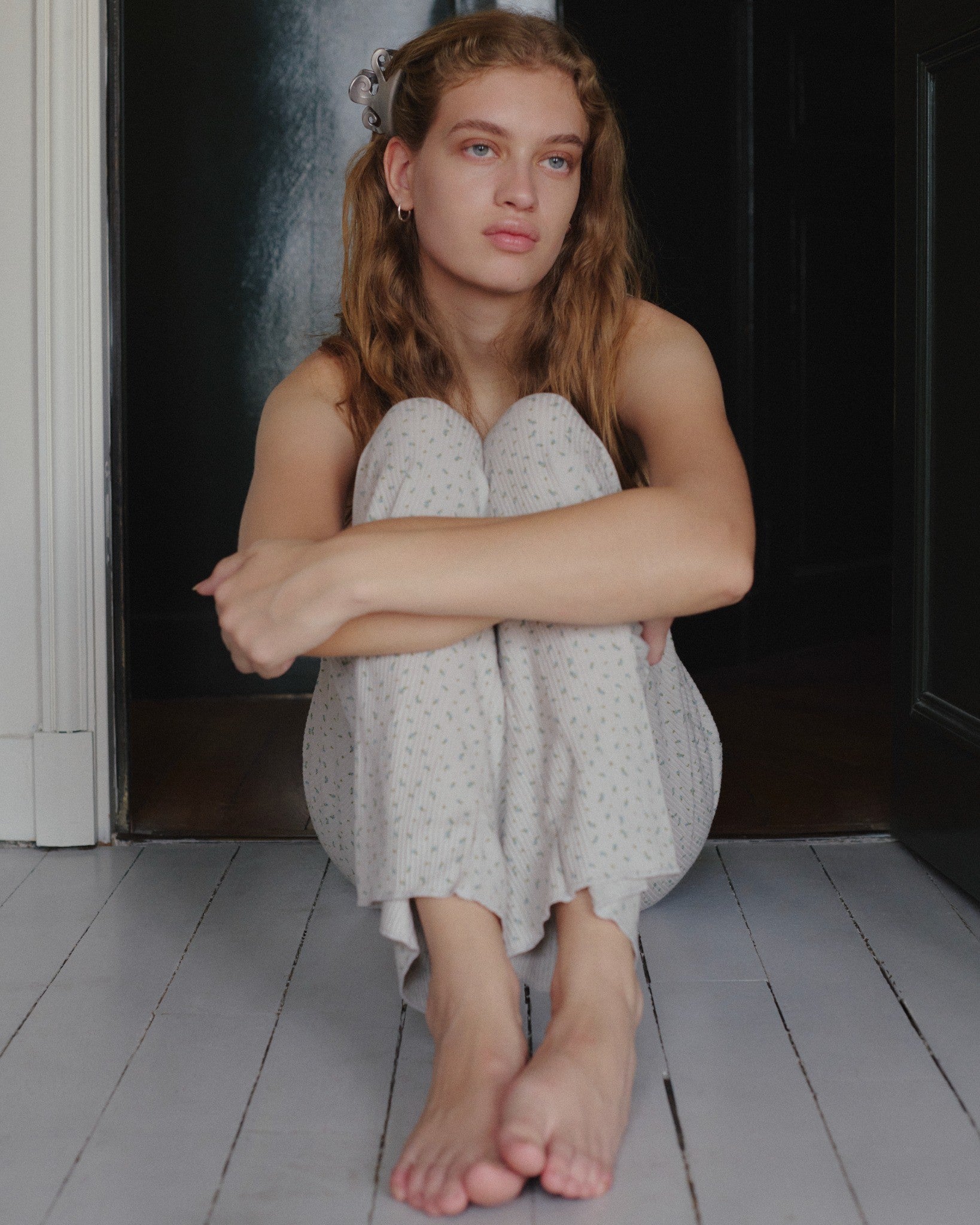 A young woman with light brown hair sits on a white wooden floor, hugging her knees to her chest. She wears the Juem Maya Pant in Blue Floral—light-colored pajamas made from comfortable, organic cotton—and looks thoughtfully into the distance.