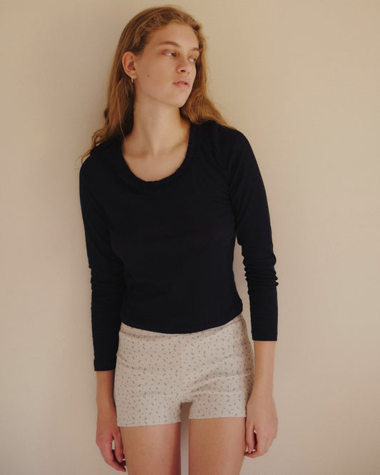 A woman with long, wavy hair wears the Claude Long Sleeve in navy by 0—an organic cotton top with a subtle scooped neckline and delicate lettuce frill trim. She pairs it with light-colored shorts featuring a small pattern, standing against a plain beige wall and gazing to her left with a neutral expression.