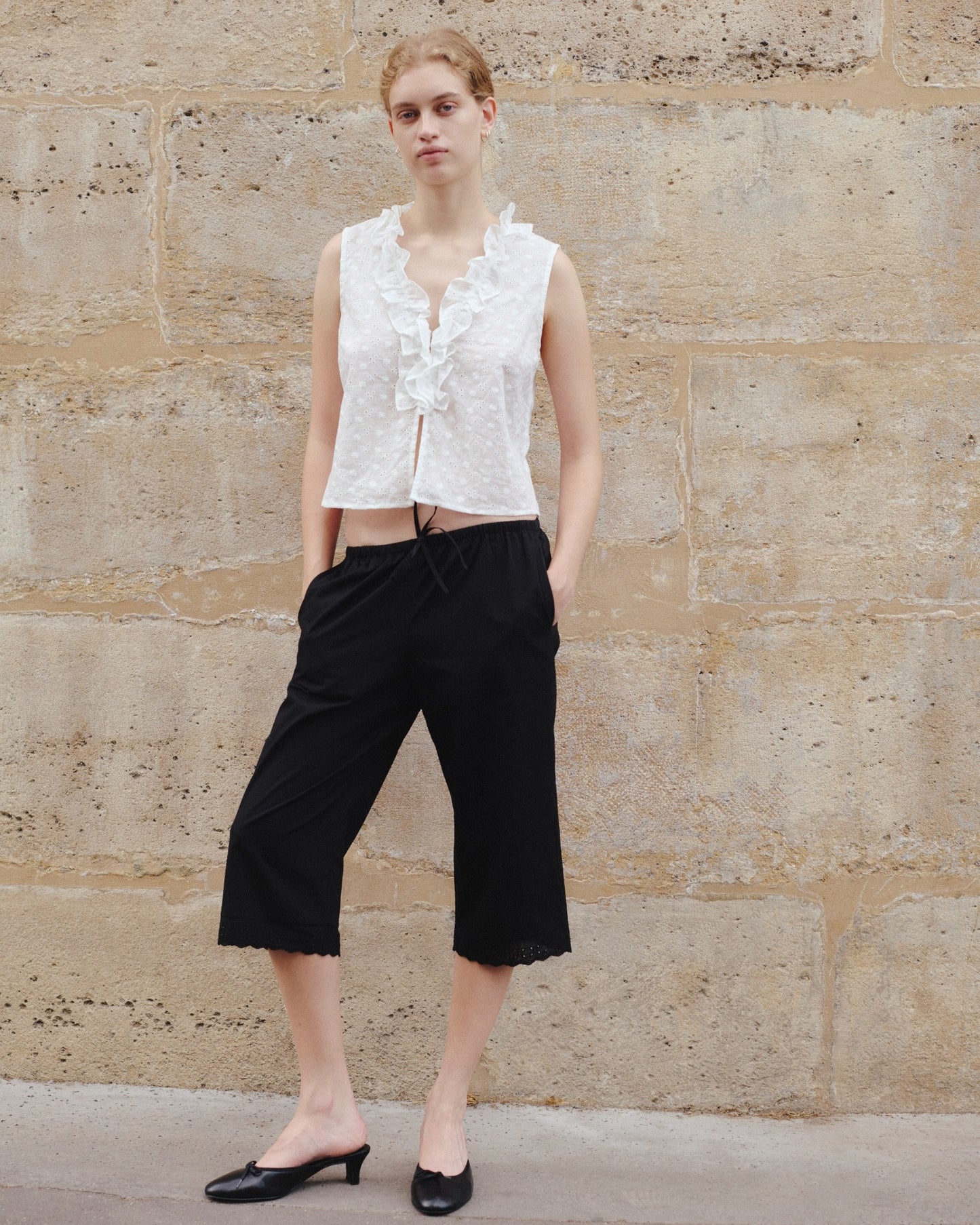 A person stands against a textured stone wall, wearing a sleeveless white ruffled top, the Ellery Capri - Black by 0, and black low-heeled shoes.