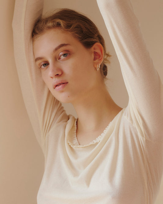 A person with light skin and wavy, light brown hair poses with arms raised, wearing the 0 Claude Long Sleeve in Butter—featuring subtle lettuce frill trim—and small hoop earrings, looking calmly at the camera against a soft beige background.