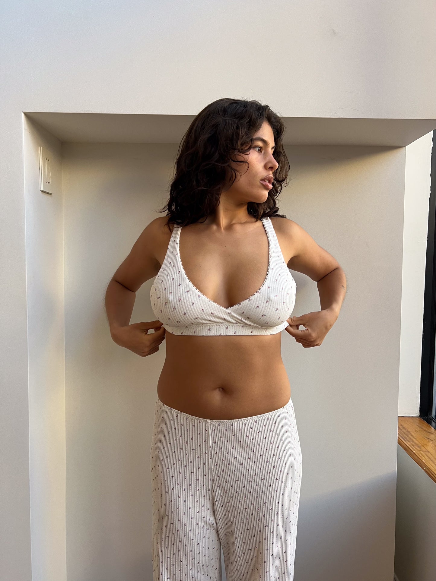 A woman with wavy brown hair stands indoors in natural light, wearing the Maya Pant - Flora and a matching bralette. She adjusts her top, enjoying the comfortable fit of her light-colored, patterned organic cotton outfit.