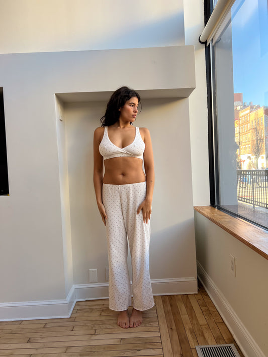 A woman in white patterned pajamas stands indoors by a sunny window. The room’s wooden floors and white walls add to the calm vibe. She enjoys relaxing mornings in her Maya Pant - Flora, made from organic cotton for a comfortable fit.