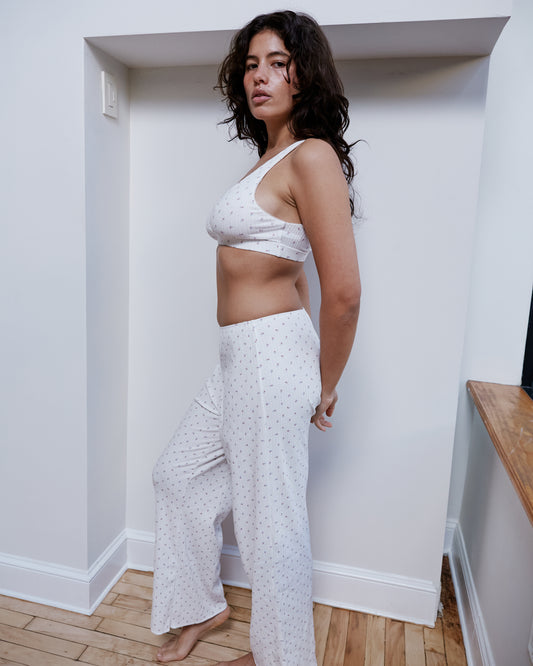 A woman with long, wavy dark hair stands indoors wearing the Bernie Bra - Flora, a matching white pajama set with small patterns and supportive underbust. She poses barefoot, sideways, against a white wall on a wooden floor.