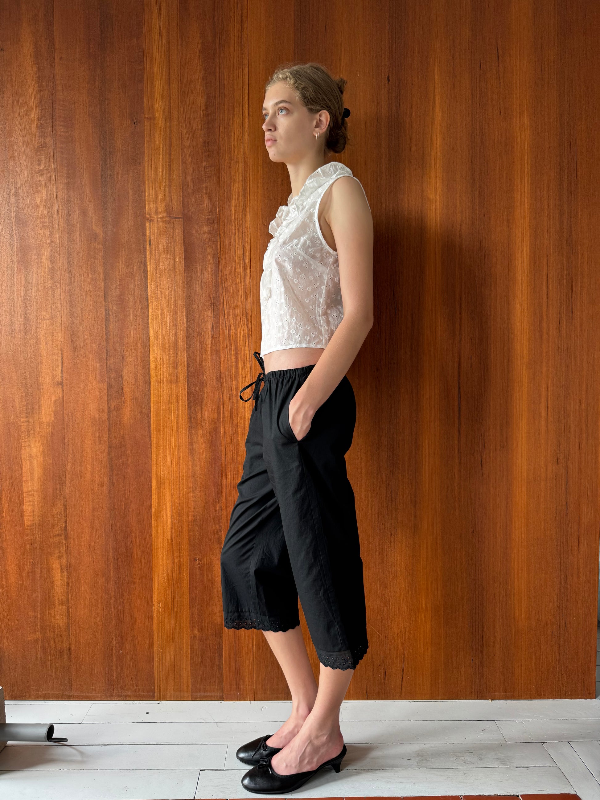 A person stands sideways against a wooden wall, wearing a white sleeveless top with delicate broderie anglaise details, the Ellery Capri - Black from 0, and black flats. They have one hand in their pocket and look forward with a neutral expression.