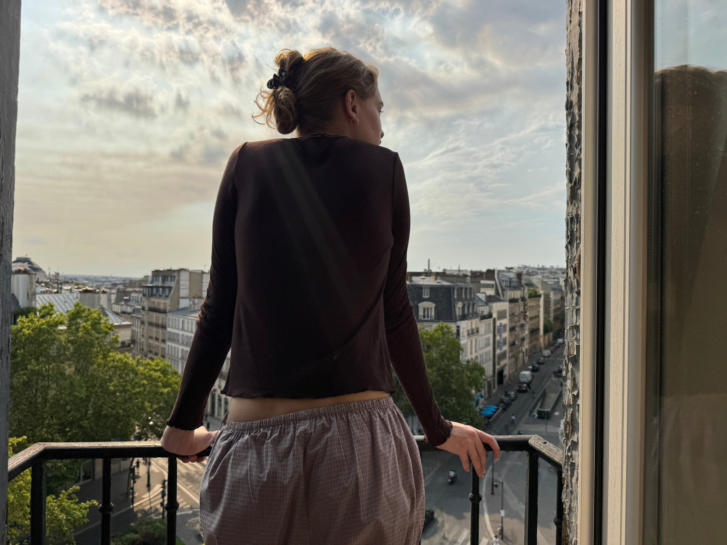 A person wearing the Rosa Long Sleeve - Chocolate by 0, featuring an organic cotton trim, stands on a balcony overlooking a city street lined with buildings and trees under a partly cloudy sky, facing away from the camera.