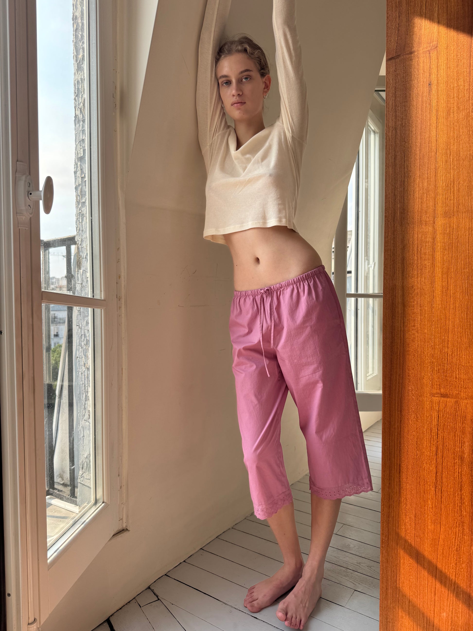 A barefoot person wearing a cream long-sleeve top and the 0 Ellery Capri - Mauve stands indoors by a window, arms raised above their head, with natural light illuminating the space.