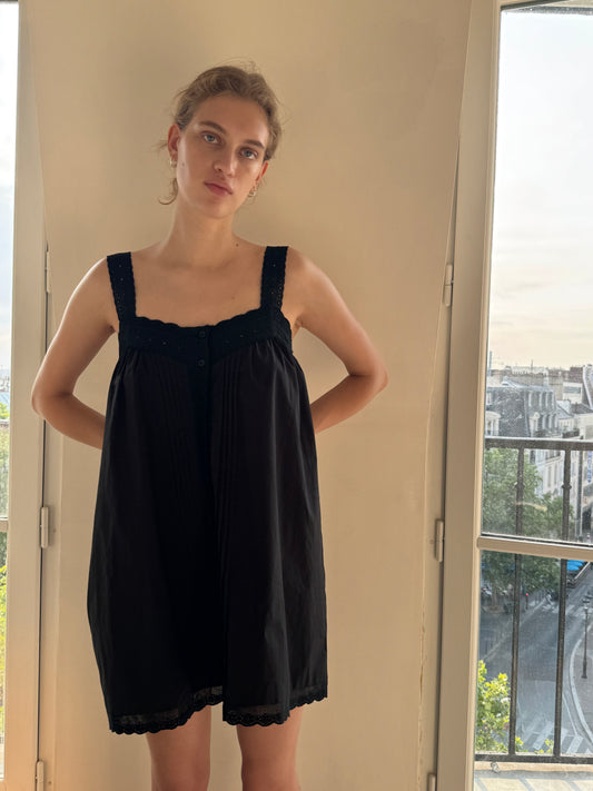 A woman wearing the Ottie Dress - Black by 0 stands indoors beside a tall window, looking at the camera. Sunlight brightens the room and city buildings are visible outside.