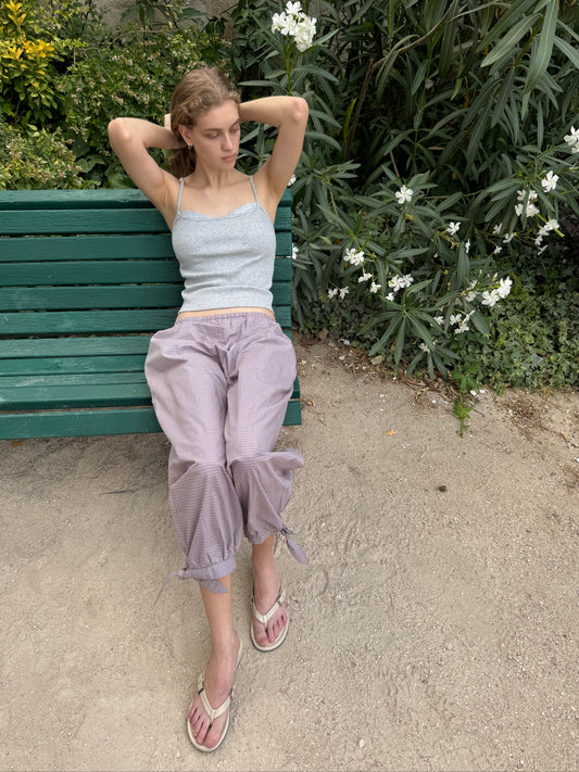 A woman wearing the 0 Joan Tank - Grey, a ribbed jersey tank with adjustable straps, and loose lavender pants sits on a green bench with her arms behind her head, looking relaxed. She pairs the look with flip-flops and is surrounded by lush greenery and blooming white flowers.