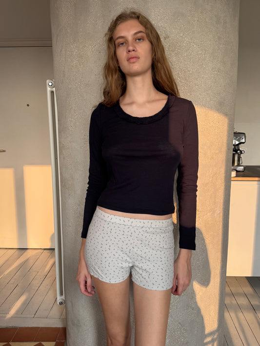 A woman with long blonde hair wearing the Claude Long Sleeve in Navy by 0 and light shorts stands indoors against a cylindrical beige pillar, bathed in warm sunlight.