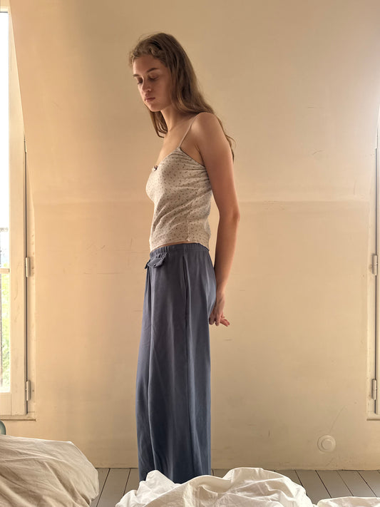 A woman with long hair stands sideways in a softly lit room, her eyes closed, wearing a light-colored tank top and the Vera Track Pant - Sea Blue by 0, featuring a relaxed fit and an elastic waistband. White bedding is visible in the foreground.