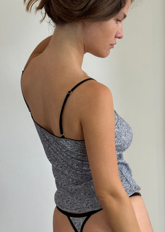 A side angle portrait of a woman holding her brown hair up in her hand, she is wearing the Harri G-string in Midnight Floral with the Margot Tank in Midnight Floral by Juem. She is looking down and standing in front of a white wall. 