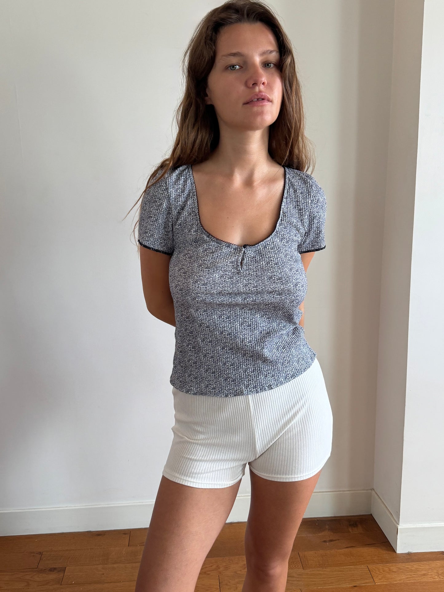 A girl is standing in front of a white wall looking into the camera with her arms crossed behind her back. She wears the Effie Tee in Midnight Floral by Juem with white shorts. She is standing on timber floorboards, and has wavy brown hair. 