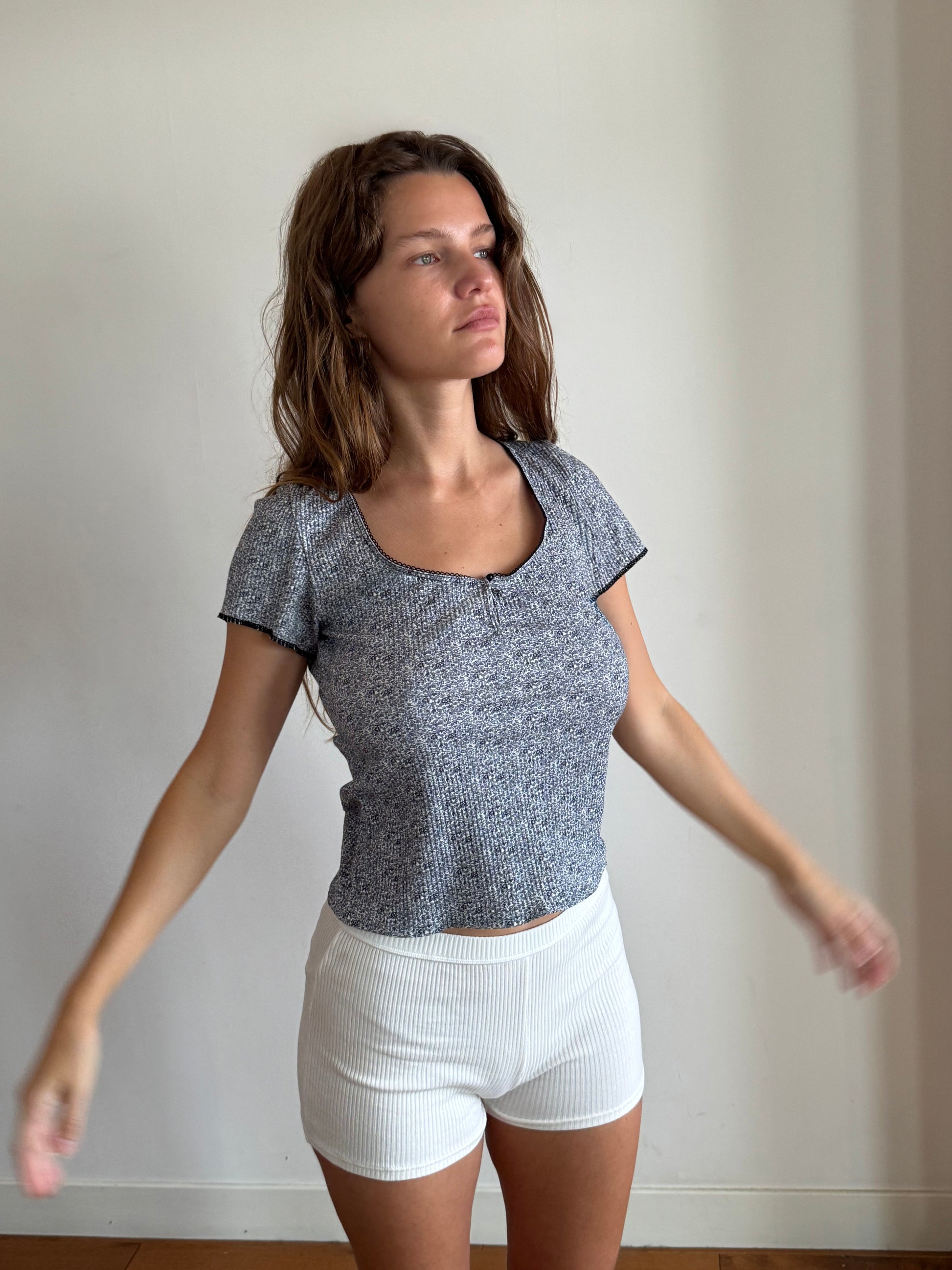 A brunette woman has her arms slightly raised, standing on floorboards in front of a white wall. She is wearing the Effie Tee in Midnight Floral by Juem with white boyleg shorts, and is looking out a window. 
