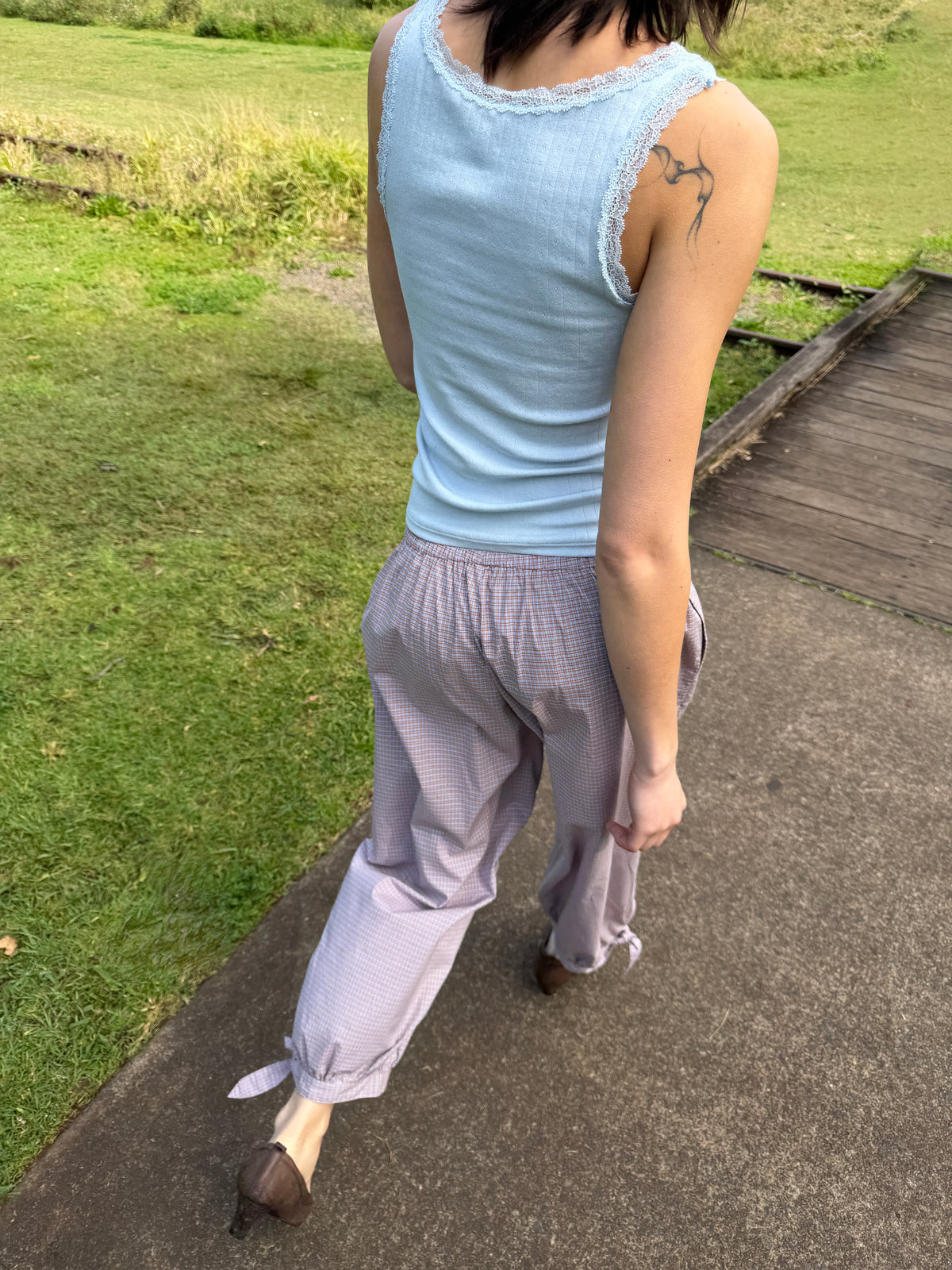 A model walks along a path through grass, seen from behind, wearing the Betty Singlet - Glacier Blue by Juem, a pointelle knit tank with lace-trimmed neckline and armholes, styled with the Freya Pant in Blue Tan Check.