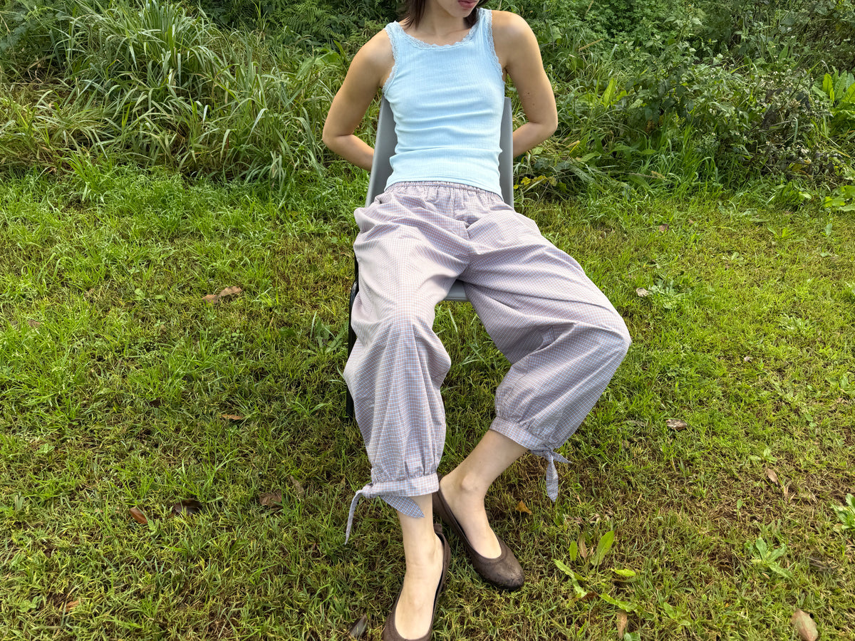 A model reclines on the grass outdoors, wearing the Betty Singlet - Glacier Blue by Juem, a pointelle knit tank with lace-trimmed neckline and armholes, styled with the Freya Pant in Blue Tan Check and brown flats.