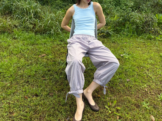 A model reclines on the grass outdoors, wearing the Betty Singlet - Glacier Blue by Juem, a pointelle knit tank with lace-trimmed neckline and armholes, styled with the Freya Pant in Blue Tan Check and brown flats.