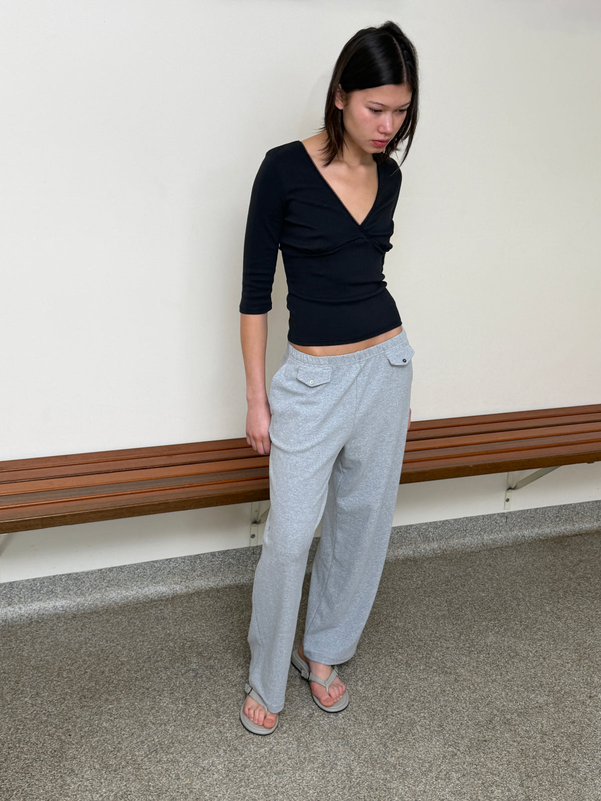 A model stands against a cream wall looking down, wearing the Fern Top - Black by Juem, a soft pointelle knit top with a v-neck crossover front and 3/4 sleeves, styled with the Vera Track Pant in Grey and sandals.
