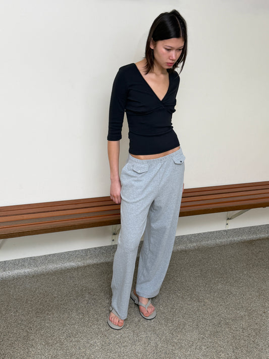 A model stands against a cream wall looking down, wearing the Fern Top - Black by Juem, a soft pointelle knit top with a v-neck crossover front and 3/4 sleeves, styled with the Vera Track Pant in Grey and sandals.