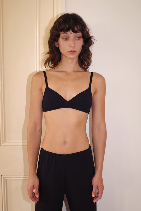A woman with wavy brown hair stands by a cream door, wearing the 0 Sophia Soft Tri - Black bra and matching black pants, looking to the side with a neutral expression.