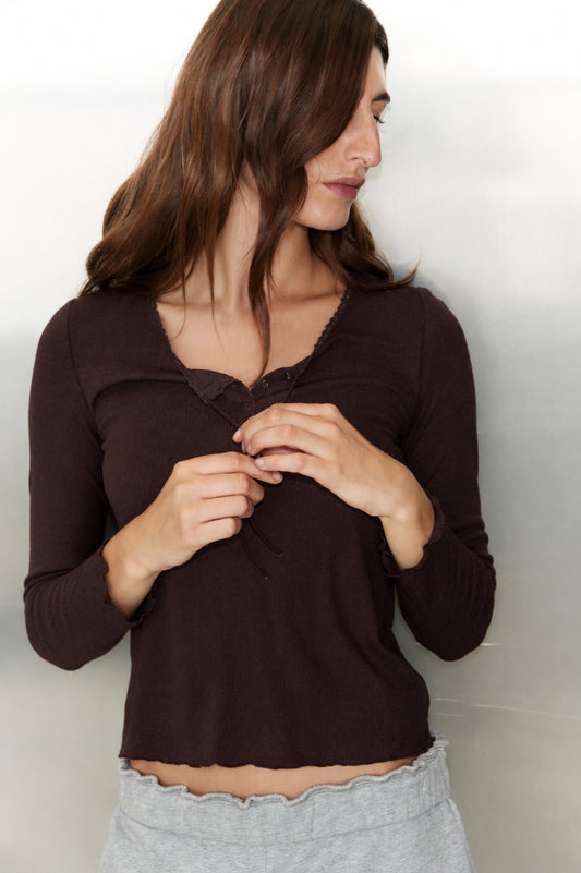 A woman with long brown hair wears the 0 Rosa Long Sleeve in Chocolate, featuring organic cotton trim, paired with light gray pants. She stands against a light background, looking left and holding her shirt’s tie near her chest.