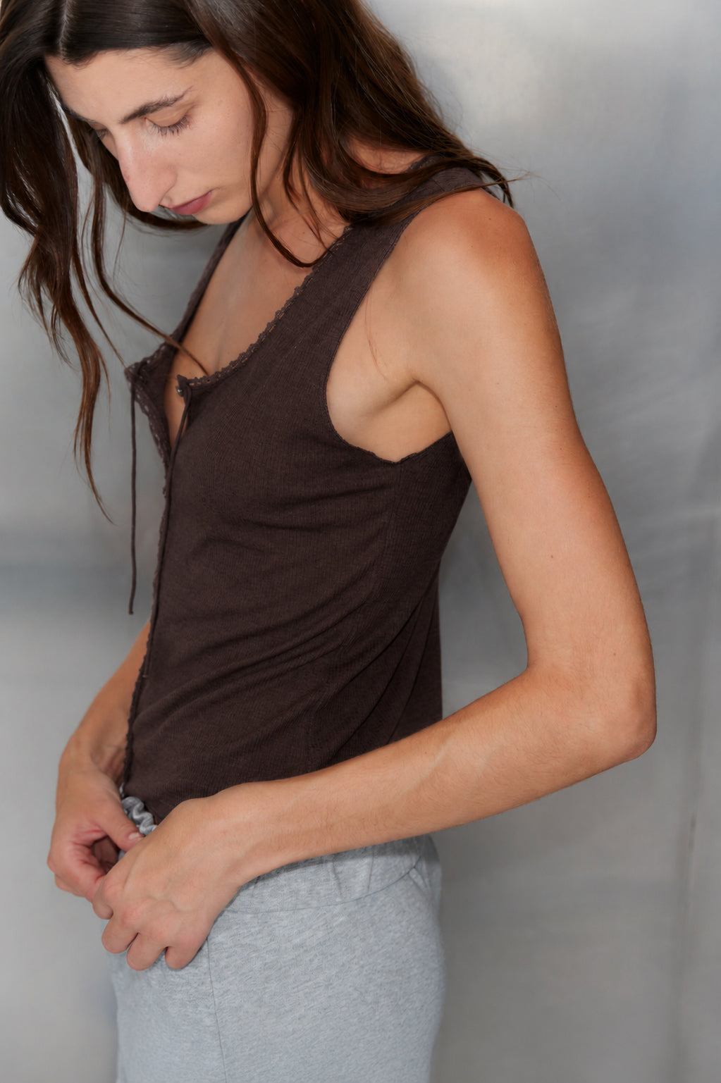 A person with long brown hair wears the Monte Tank - Chocolate by 0, a sleeveless modal-linen top with a scoop neckline, paired with light gray pants, and adjusts their waistband against a plain, neutral background.