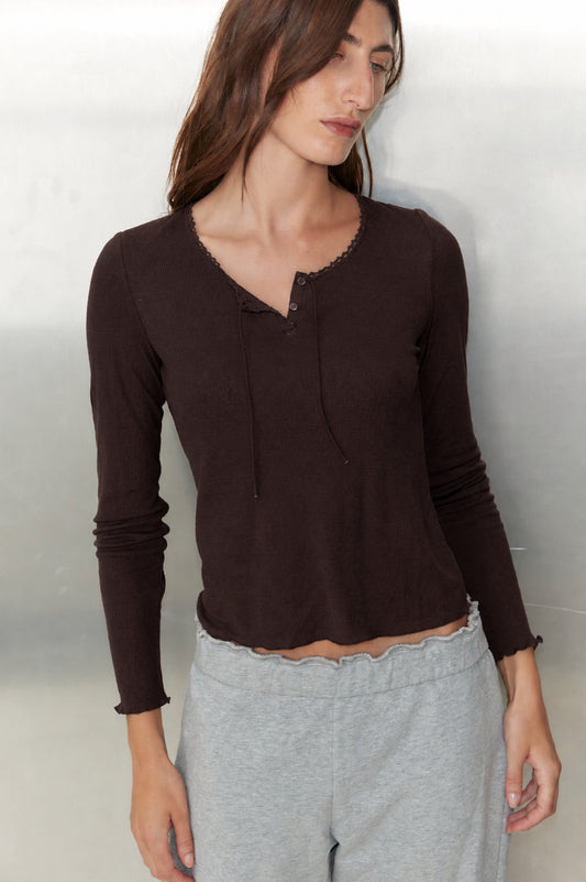 A woman with long brown hair wears the Rosa Long Sleeve - Chocolate by 0 and light gray sweatpants, standing against a light metallic background and looking slightly downward.